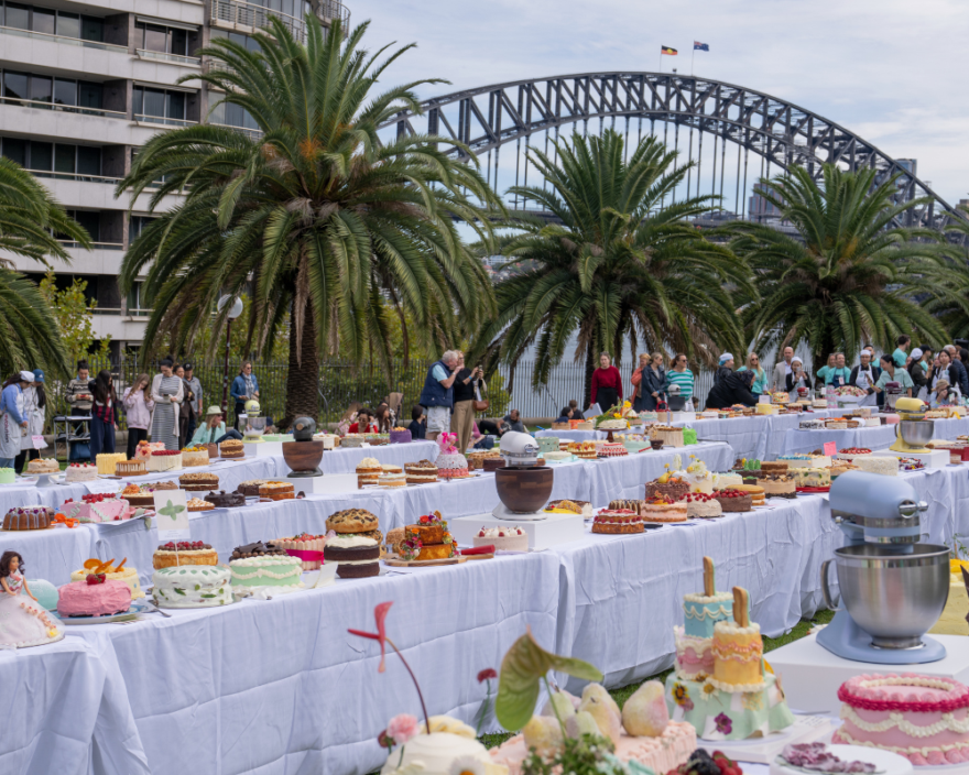 Outdoor cake festival with dozens of decorated cakes on white-clothed tables, palm trees, and a steel arch bridge in the background.