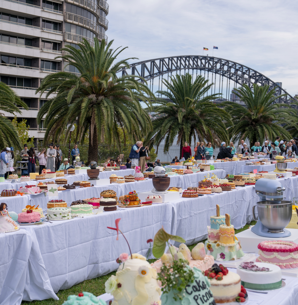 Outdoor cake festival with dozens of decorated cakes on white-clothed tables, palm trees, and a steel arch bridge in the background.