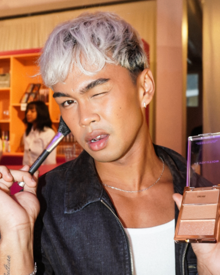 Person applying makeup with a brush while holding a compact powder palette, winking at the camera in a bright room