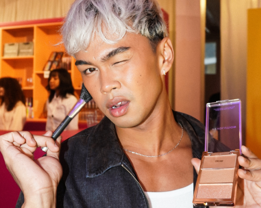 Person applying makeup with a brush while holding a compact powder palette, winking at the camera in a bright room
