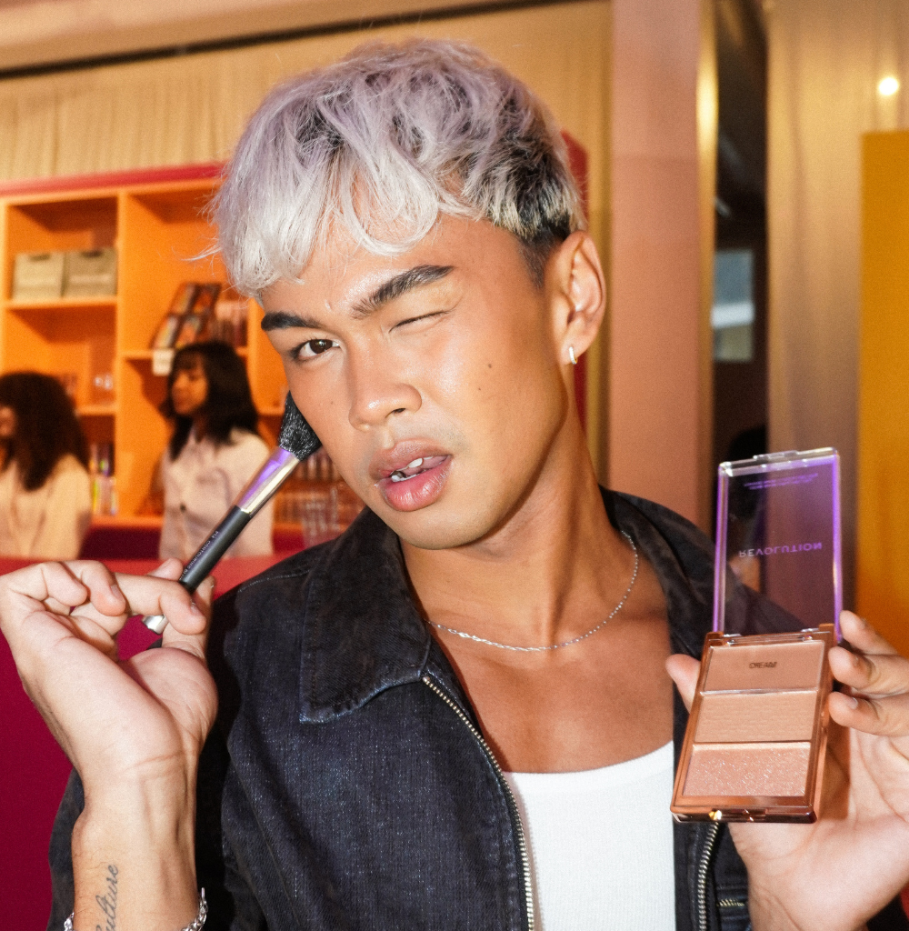 Person applying makeup with a brush while holding a compact powder palette, winking at the camera in a bright room