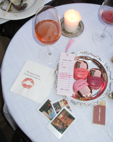 Table setting with a lit candle, two wine glasses, and a silver tray displaying pink nude by nature makeup items.