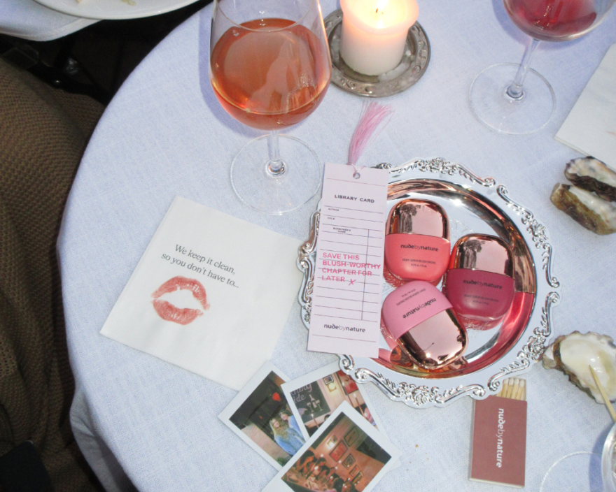 Table setting with a lit candle, two wine glasses, and a silver tray displaying pink nude by nature makeup items.