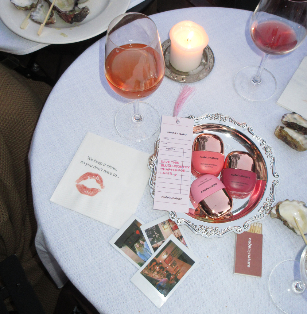 Table setting with a lit candle, two wine glasses, and a silver tray displaying pink nude by nature makeup items.
