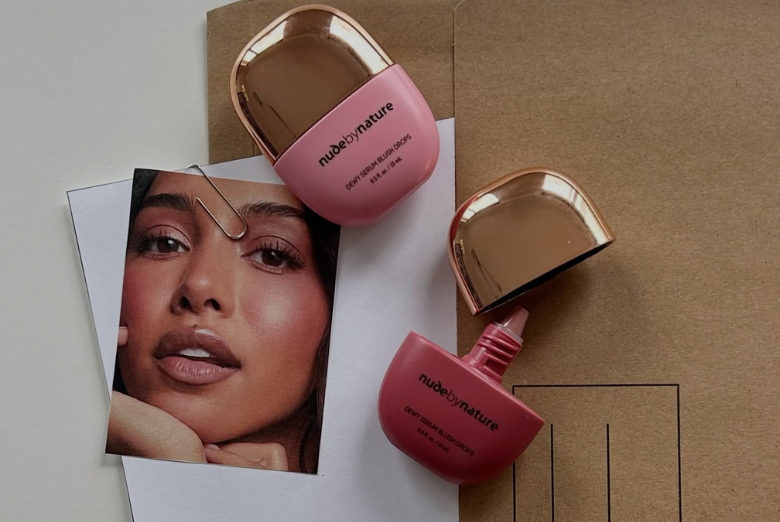 Two nude by nature makeup bottles with rose-gold caps on a light surface beside a model’s photo on white paper.
