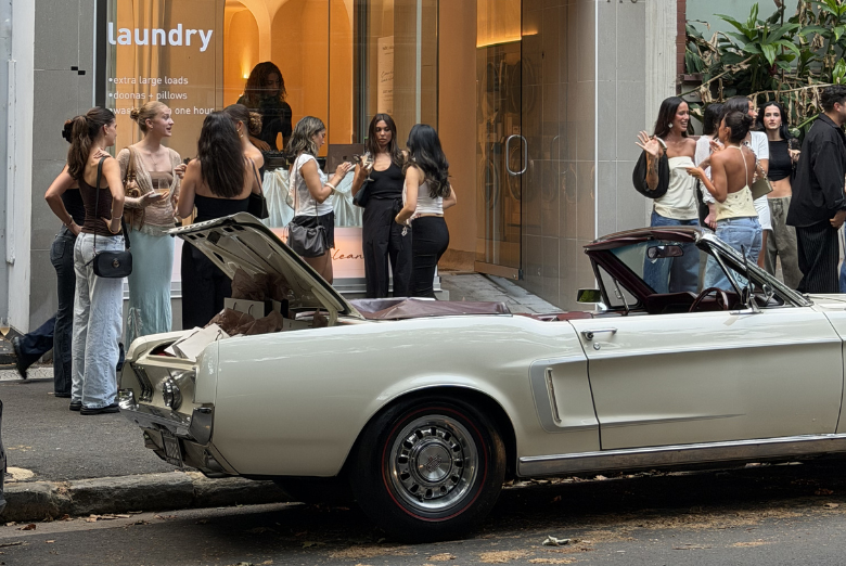 Group of people chat near an open-top white classic car parked on a city street in front of a shop with a laundry sign.
