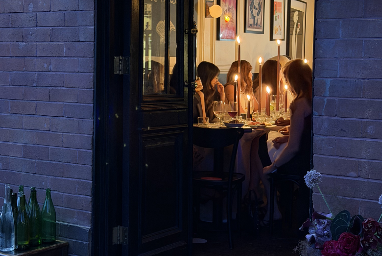 Group of people dining by candlelight inside a restaurant, viewed through an open door from outside.
