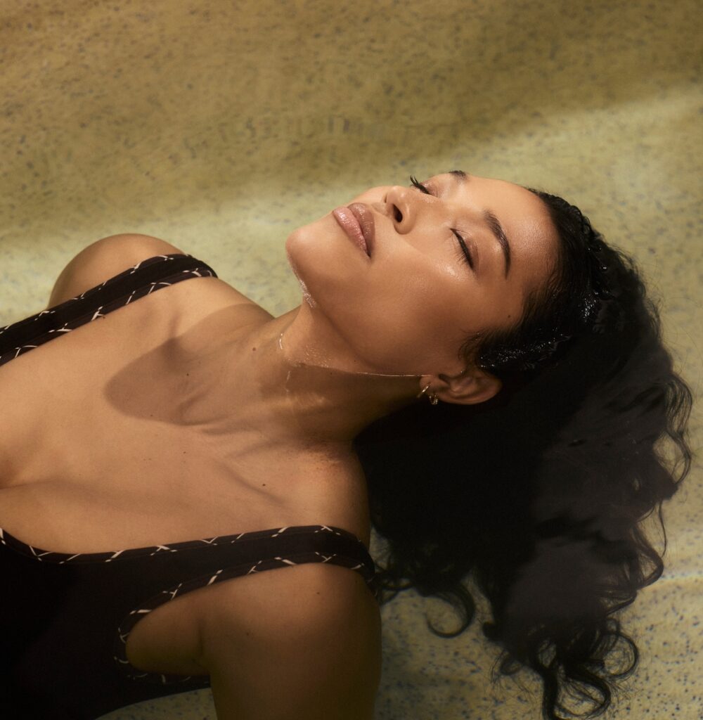 Woman floating in a shallow pool with eyes closed, wearing a black swimsuit with white stitching detail.