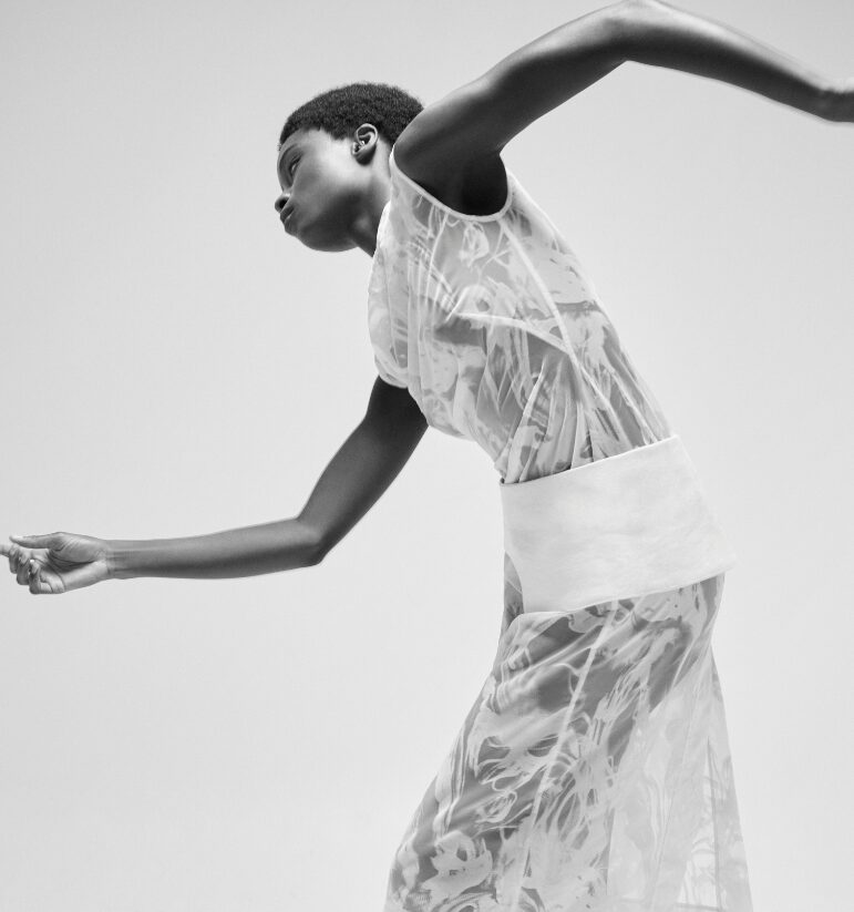 Black-and-white fashion photo of a person in a sheer patterned dress, arms extended in a dynamic mid-motion pose against a plain background.