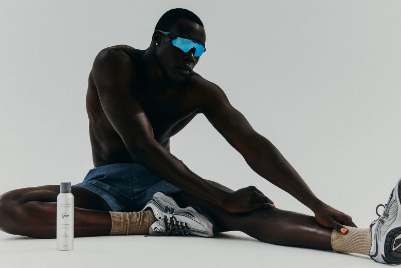 Athletic Black man in blue shorts sits on a white studio floor, stretching his legs, wearing blue mirrored sunglasses; a grooming product bottle stands nearby.