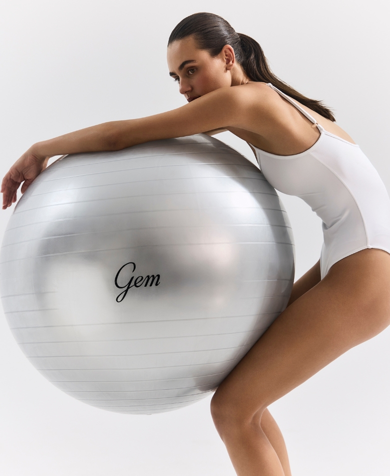 Woman in a white leotard leaning on a large silver exercise ball labeled 'Gem' during a fitness pose.