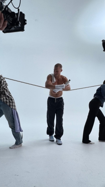 Shirtless man on a white studio set, gripping a rope as crew members and cameras surround him (behind the scenes).