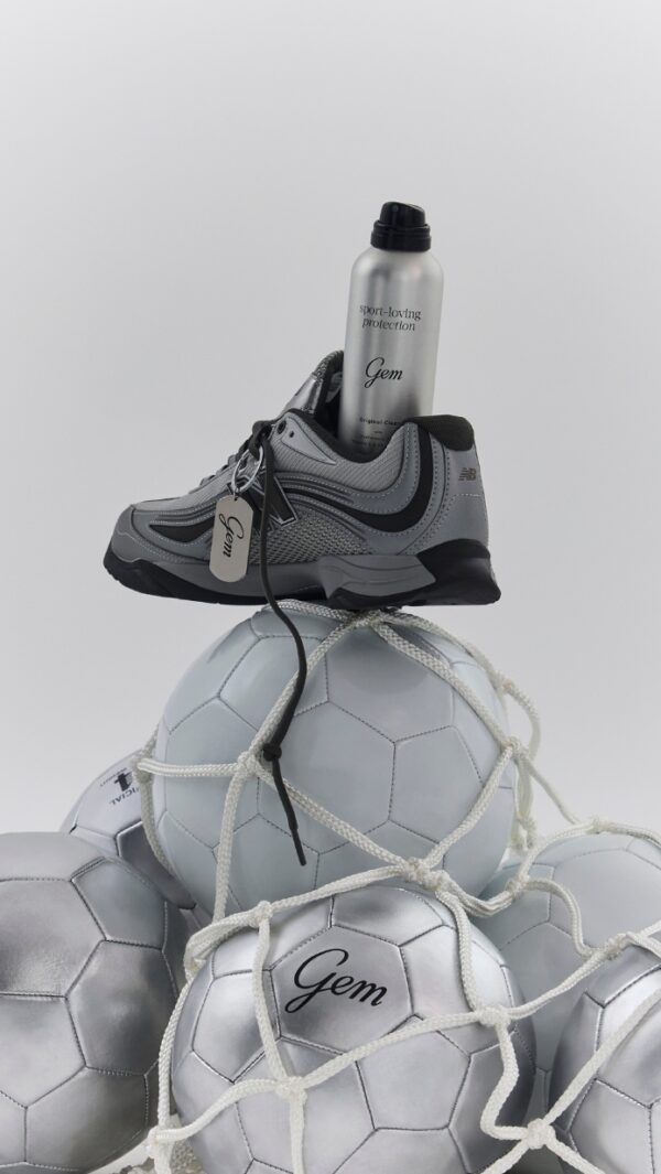 Gray athletic sneaker perched on a stack of silver soccer balls tied with white netting, with a silver spray bottle labeled Gem tucked into the shoe.