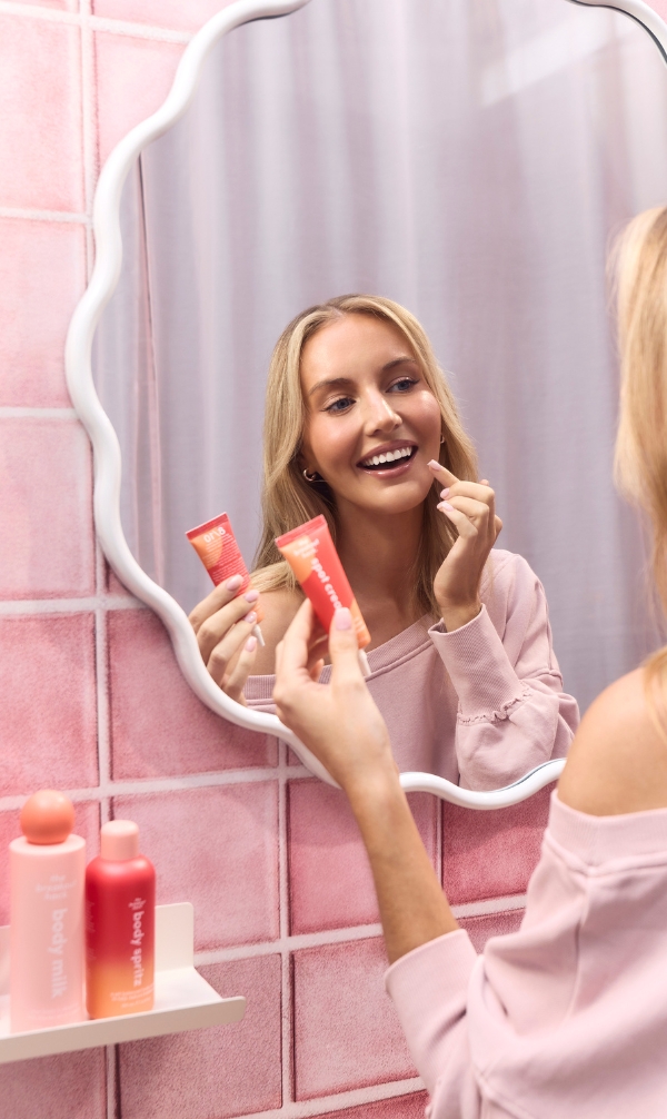 Woman in a pink sweater applies lip product in a pink-tiled bathroom, looking into a wavy-mirrored mirror while holding two cosmetic tubes.