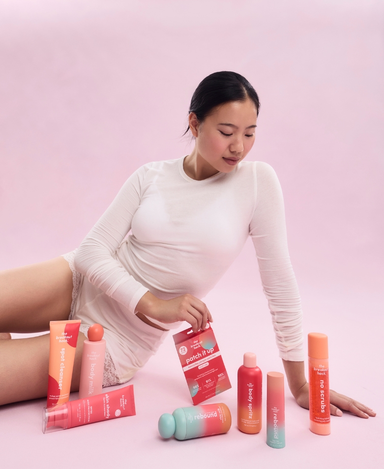 Asian woman in white sits on a pink backdrop surrounded by pastel skincare bottles and a pink card.