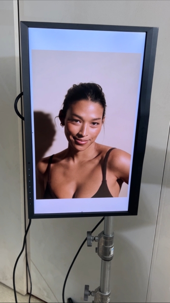 Portrait of a smiling woman in a black tank top, displayed on a mounted screen on a stand in a studio setting.