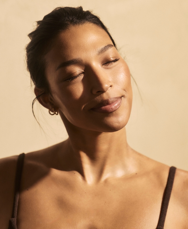 Woman with eyes closed and a serene smile, basking in warm sunlight against a beige backdrop.