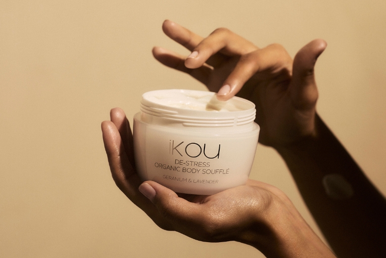 Hands cradle a white jar of ikou Organic Body Soufflé (Geranium & Lavender) with cream being scooped from the lid.