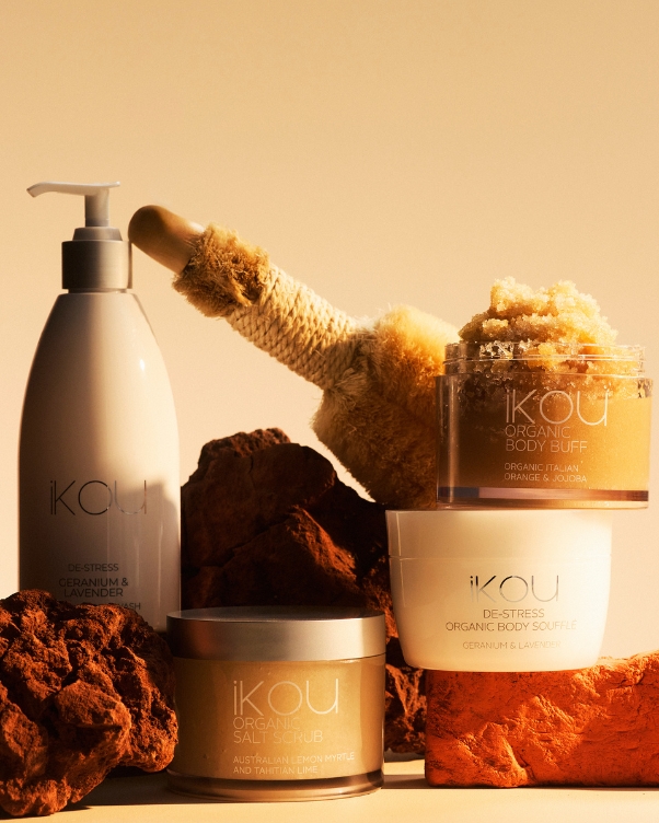 IKOU organic body care collection displayed on rocky, warm-toned backdrop with a body scrub jar and a pump bottle nearby.