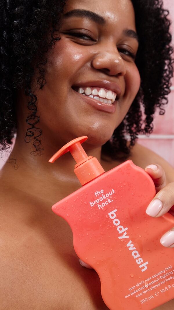 Smiling woman holds a coral-pink pump bottle labeled 'the breakout hack' body wash: close-up portrait with product