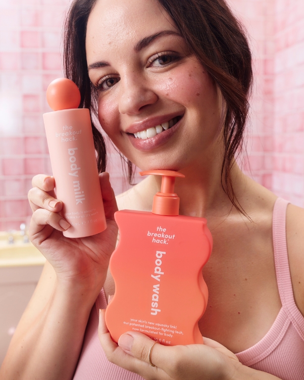 Smiling woman holds two pink skincare bottles: body wash and body milk, in a pink bathroom setting.