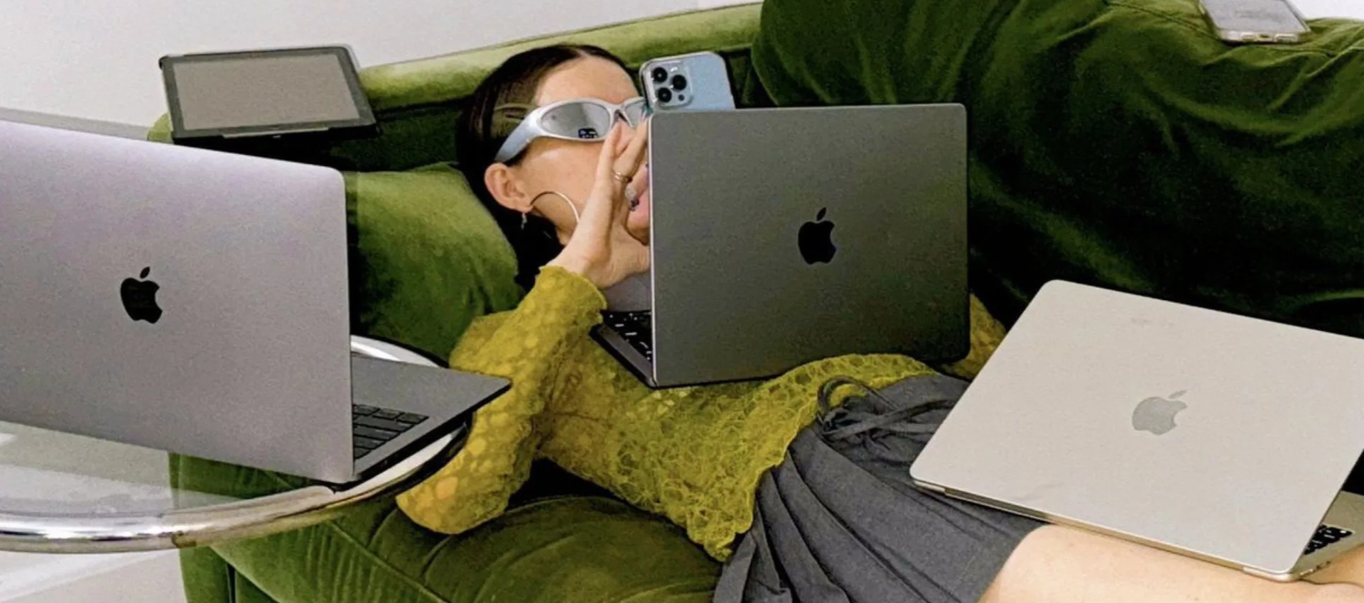 Person wearing white protective goggles sits on a green velvet sofa surrounded by three Apple laptops and a blue iPhone nearby.