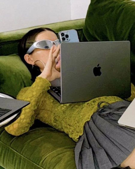 Person wearing white protective goggles sits on a green velvet sofa surrounded by three Apple laptops and a blue iPhone nearby.
