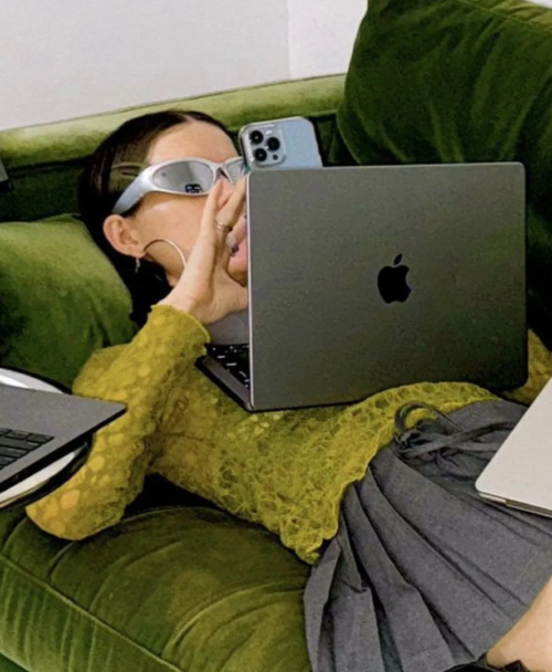 Person wearing white protective goggles sits on a green velvet sofa surrounded by three Apple laptops and a blue iPhone nearby.