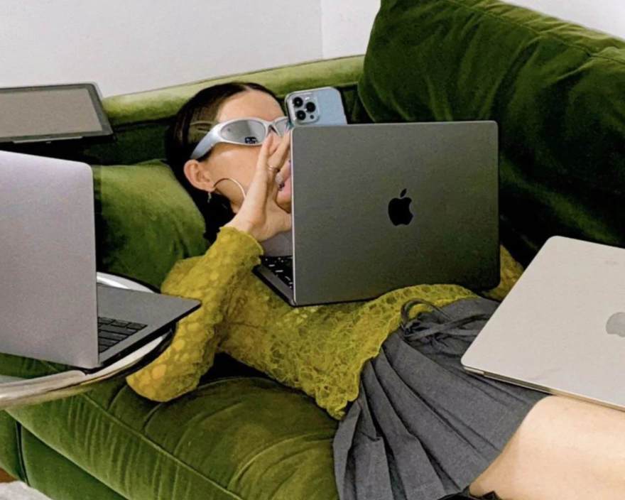 Person wearing white protective goggles sits on a green velvet sofa surrounded by three Apple laptops and a blue iPhone nearby.